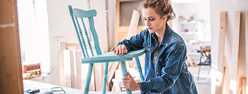 Woman refinishing a blue wooden chair in her shop.